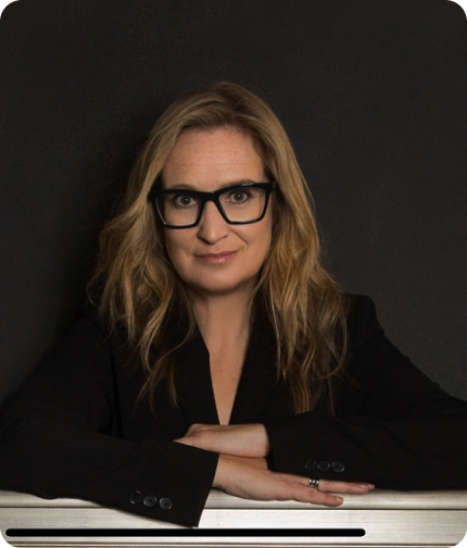 Image shows a blond women in a business suit, wearing black-framed glasses